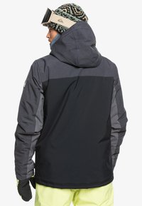 Herrenjacke in Schwarz und Grau mit Kapuze, mit einer strukturierten Oberfläche und kontrastierenden Einsätzen, kombiniert mit einer gemusterten Strickmütze.