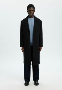 Zwarte wollen overjas met een notch-kraag, een eenrijige ontwerp en grote knopen, gelaagd over een lichtblauw geribbeld sweater en donkere broeken.