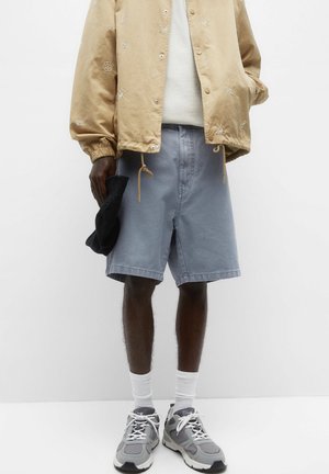 Light beige embroidered bomber jacket, white sweater underneath, light blue shorts, and gray sneakers with white socks. Held black item in hand.