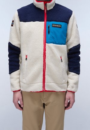 Mand iført fleecejakke med cremet, marineblå og blå paneler, rød lynlåskant og beige bukser mod en ensfarvet baggrund.