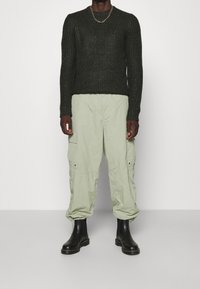 Dunkelgrüner gestrickter Pullover mit gerippter Textur, hellgrüne Cargo-Shorts und schwarze Knöchelstiefel. Einfache goldene Kettenhalskette als Akzent.