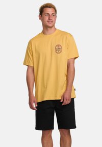 Geel katoenen t-shirt met korte mouwen, ronde hals en een rond logo op de borst. Gecombineerd met zwarte shorts. Gladde textuur.