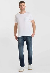 Witte t-shirt met korte mouwen en een borstzakje, gecombineerd met donkerblauwe spijkerbroek en witte sneakers, staand tegen een neutrale achtergrond.
