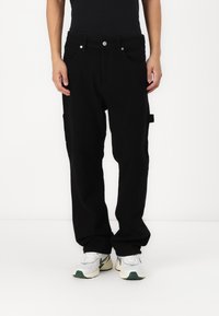Persona con pantalones negros holgados con bolsillos y zapatillas blancas de pie contra un fondo blanco liso.