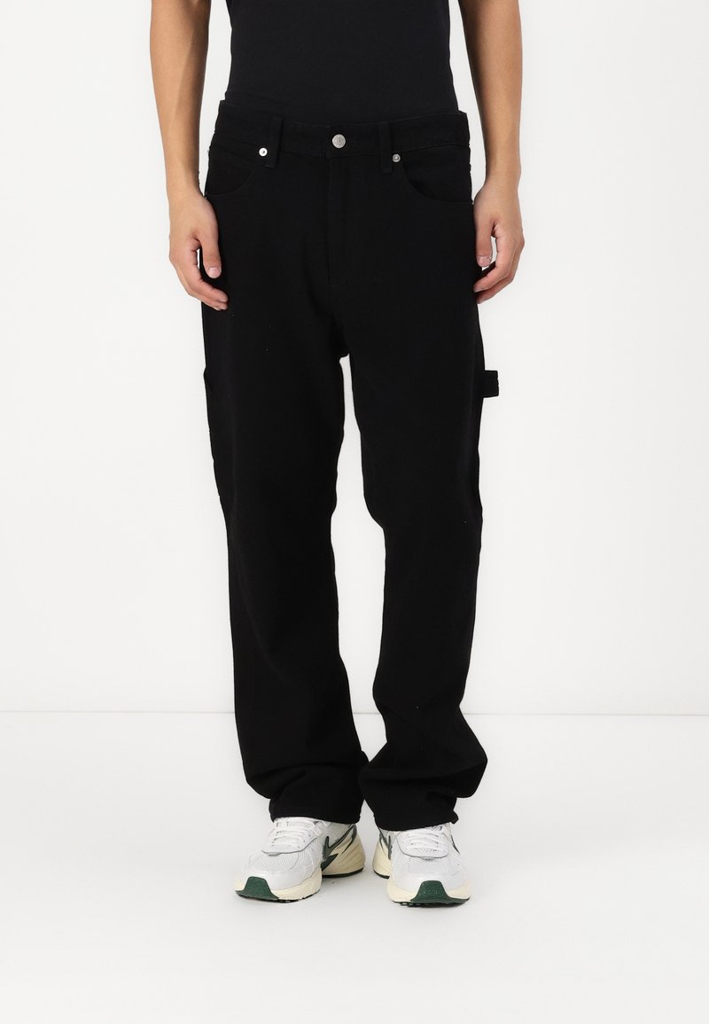 Persona con pantalones negros holgados con bolsillos y zapatillas blancas de pie contra un fondo blanco liso.