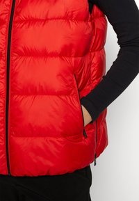 Personne portant un gilet matelassé rouge vif avec des poches zippées par-dessus une chemise noire à manches longues, la main dans la poche droite.