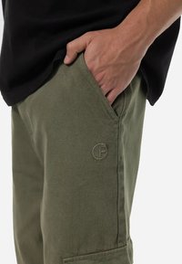 Pantaloni de culoare verde măsliniu, dintr-un material texturat, cu un logo cusut pe buzunar și o croială lejeră, dotati cu buzunare laterale.