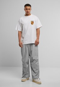 Hvid bomulds t-shirt med et lille rødt og gult våbenskjold, parret med løse grå cargo bukser og beige sneakers med elastiske manchetter.