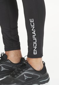 Närbild av underben som bär svarta leggings med texten "ENDURANCE" och svarta träningsskor med grå logotyp, stående på vit golv.