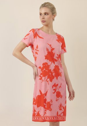 Frau trägt ein knielanges rosa Kleid mit großen roten Blumenmustern, steht mit einer Hand in der Hüfte und schaut zur Seite vor einem beigen Hintergrund.