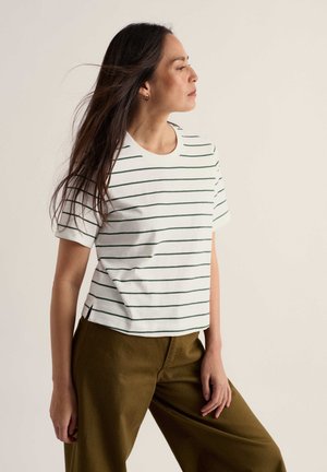Femme debout regardant sur le côté, portant une chemise blanche à manches courtes rayée et un pantalon vert olive sur un fond uni.