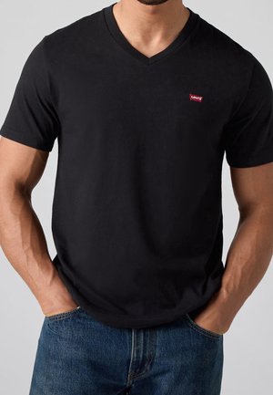 Homme portant un T-shirt Levi's noir col en V à manches courtes et un jean bleu, mains dans les poches, vue de la taille vers le haut.