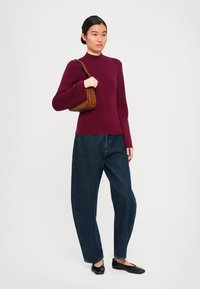 Egy nő bordó hosszú ujjú felsőt, laza sötét farmernadrágot, fekete lapos cipőt visel, és egy barna válltáskát tart egy egyszínű háttér előtt.