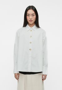 Camisa blanca de botón con tejido liso, con un cuello clásico y cinco botones circulares dorados en la parte delantera. Mangas largas, corte holgado.