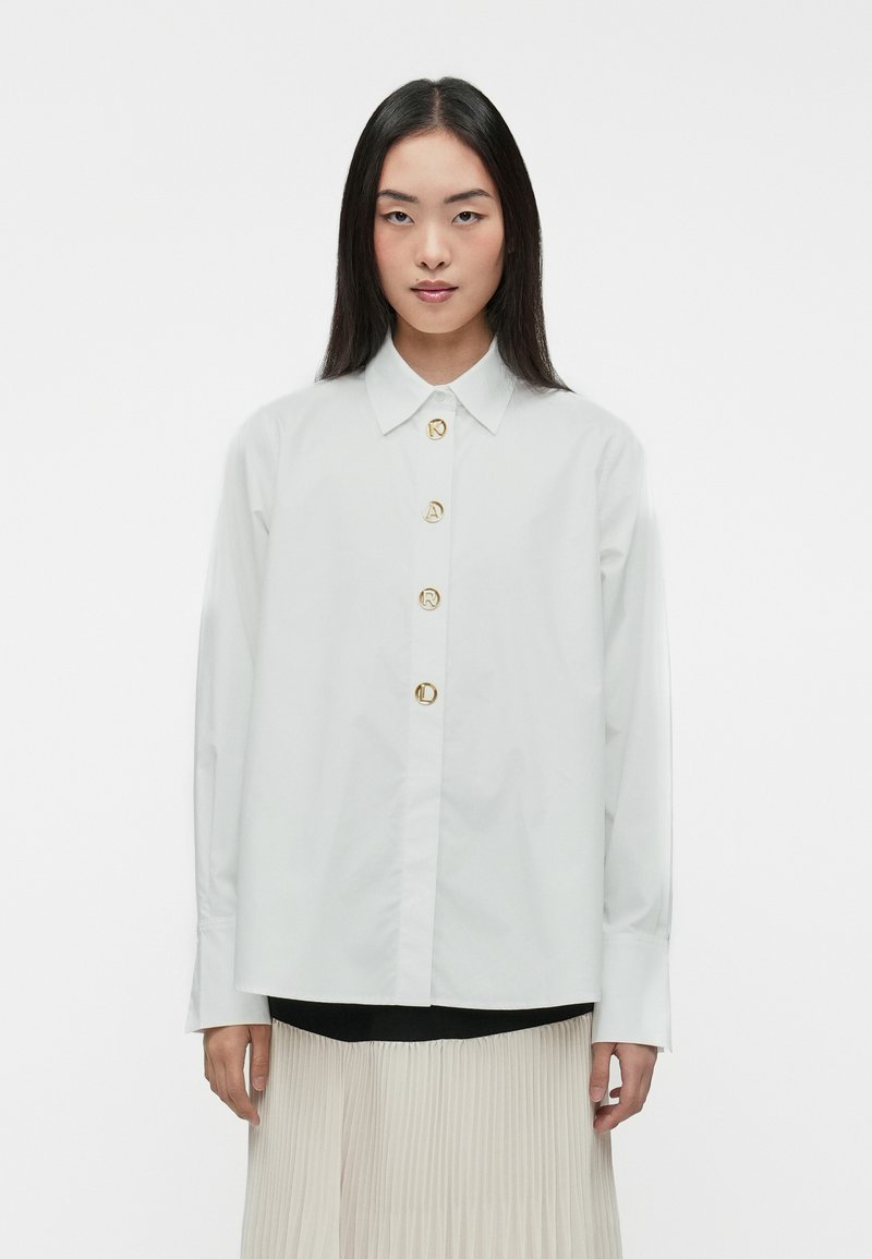Camisa blanca de botón con tejido liso, con un cuello clásico y cinco botones circulares dorados en la parte delantera. Mangas largas, corte holgado.