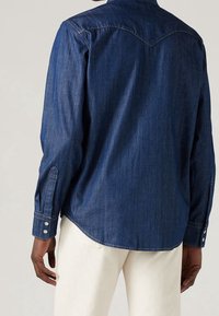 Donkerblauwe denim shirt met lange mouwen, knoopmanchetten, puntige schouderpas en een gebogen onderkant, voorzien van subtiele stikdetails.
