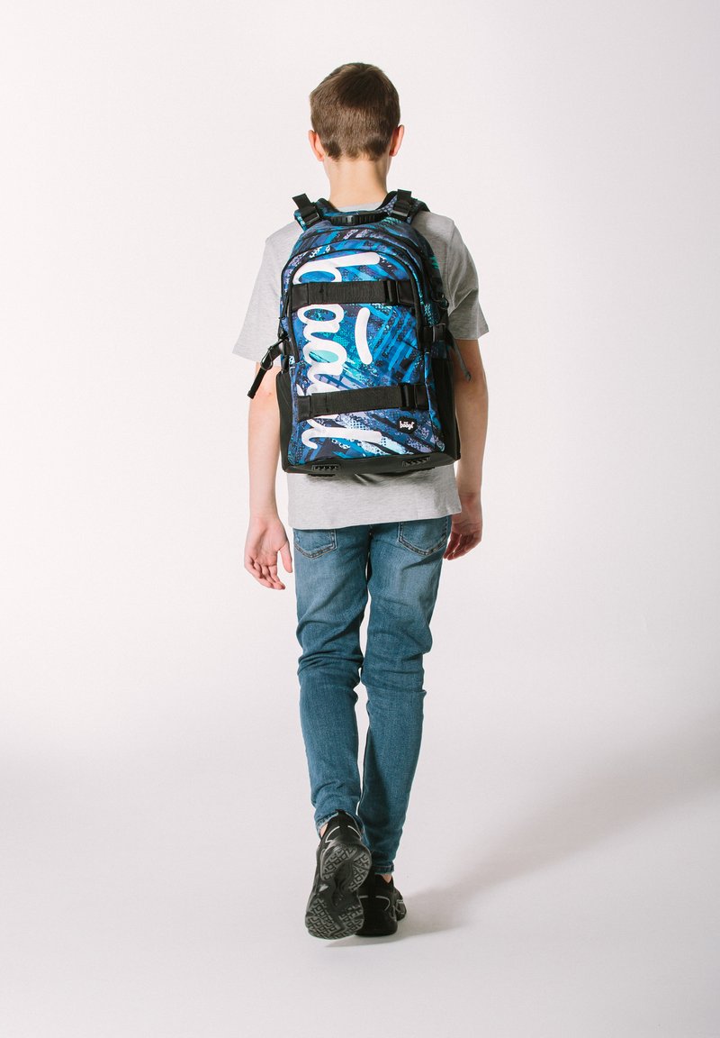 Zaino con una base nera caratterizzato da motivi astratti blu e bianchi, spallacci regolabili e molteplici tasche. Indossato da un bambino in jeans.