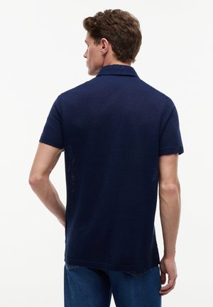 Άνδρας με κοντά σγουρά μαλλιά, φοράει μπλε κοντομάνικο πουκάμισο με γιακά και μπλε τζιν, εμφανιζόμενος από πίσω.