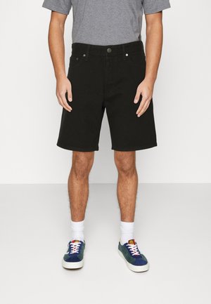 Uomo con pantaloncini neri fino al ginocchio, maglietta grigia, calzini bianchi e scarpe da ginnastica blu, in piedi su pavimento e sfondo bianco uniforme.