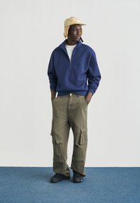 Φλις με φερμουάρ σε ναυτικό μπλε, χακί παντελόνι cargo με πολλές τσέπες, μαύρα παπούτσια και μπεζ καπέλο τύπου κάπα. Απλός, λειτουργικός σχεδιασμός.