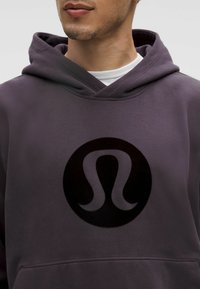 Lila bolyhos kapucnis pulóver, amelyen egy kiemelkedő fekete logó található kör alakú dizájnnal. Texturált anyag, húzós kapucnival és elülső zsebbel.