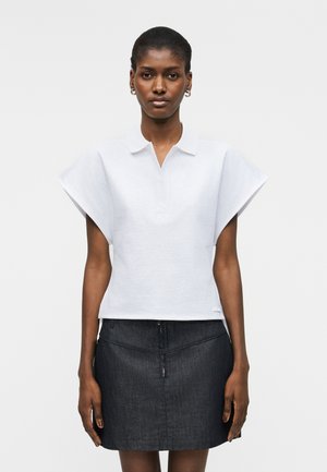 Γυναίκα που φοράει λευκό, δομημένο τοπ με κοντά μανίκια και γιακά, και σκουρόχρωμη μίνι φούστα από τζιν με ψηλή μέση, στέκεται μπροστά σε απλό φόντο.