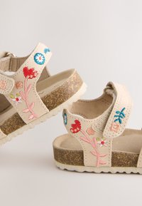 Beige sandalen met bloemrijke borduursels, verstelbare band, kurken voetbed en een witte rubberen zool met structuur. Voorzien van roze, rode en blauwe accenten.