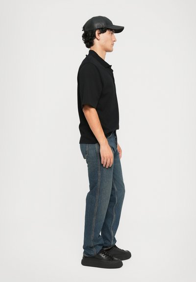 Polo shirt noir, jeans en denim bleu avec des coutures visibles, et baskets noires. Le sujet porte une casquette noire, vu de profil.