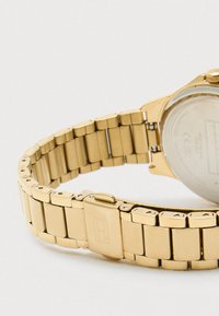 Χρυσό μεταλλικό ρολόι με βραχιόλι αλυσίδας, με φινίρισμα βουρτσισμένου μετάλλου, ορθογώνιους συνδέσμους και κλειστό σύστημα, τονίζοντας τον λεπτό, κομψό σχεδιασμό.