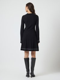 Uma mulher vestindo um vestido preto de mangas longas com textura canelada e saia plissada, com detalhes em branco, combinado com botas pretas de salto alto até o joelho, de costas.