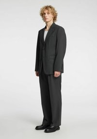Czarna marynarka z wyraźnie zarysowanymi ramionami, zapinana na jeden guzik, z klapami z wycięciem, zestawiona z pasującymi spodniami i czarnymi butami.