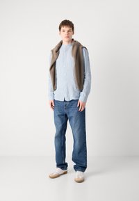 Hellblaues Langarmhemd mit Knöpfen, beiger Pullover über die Schultern gelegt, blaue Jeans und weiße Turnschuhe mit hellen Sohlen.