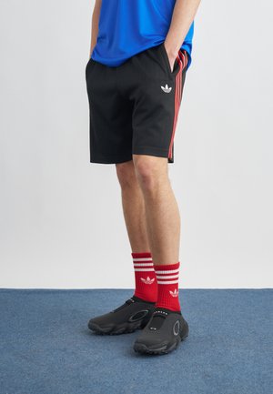 Άνδρας που φοράει μαύρα σορτς Adidas με κόκκινες ρίγες, κόκκινες κάλτσες Adidas, μαύρα παπούτσια Oakley slip-on και μπλε πουκάμισο, στέκεται πάνω σε μπλε χαλί.