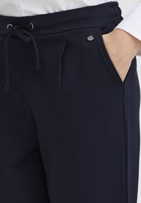 Pantaloni de trening bleumarin cu talie elastică și șnur reglabil, prevăzuți cu buzunare laterale, un mic logo metalic rotund și textură netedă.