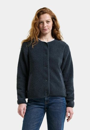 Kvinde med langt bølget rødt hår iført en mørkegrå fleecejakke med trykknapper og blå jeans, stående mod en ensfarvet lys baggrund.