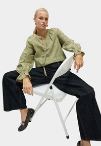 Olijfgroene geborduurde blouse met zwierige mouwen, gecombineerd met donkere denim wijdvallende jeans en bruine ballerina's. Eenvoudige striksluiting.