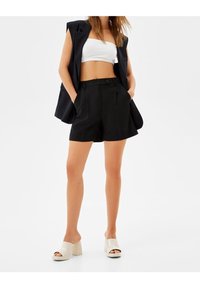 Svarta skräddarsydda shorts med två framfickor, hög midja, veckad design och knappstängning. Kombineras med en vit kort topp och beiga klackskor.