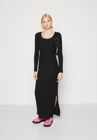 Vestido maxi de malha canelada preto com mangas longas, possui uma fenda lateral. Combinado com botas chunky rosas brilhantes e uma pequena bolsa preta.