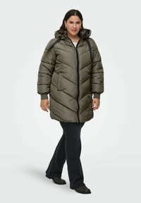 Padded olive green coat with a hood, featuring a zip front, chevron stitching, and side pockets. Worn over black pants and shoes.