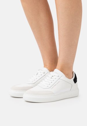 Paire de baskets basses blanches avec lacets, détail beige clair sur le bout, patch noir au talon, et logo Calvin Klein Jeans sur le côté et la semelle.