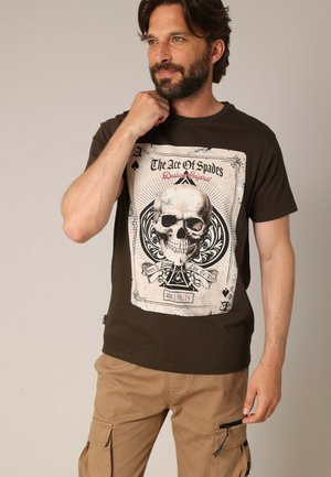 T-shirt en coton marron avec un grand design de crâne et de carte à jouer en noir et blanc, avec le texte "L'As de Piques" au-dessus et une iconographie en dessous.