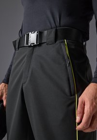 Pantalones negros de material suave con corte Slim, que cuentan con un llamativo detalle de cremallera amarilla y un cinturón negro resistente con hebilla metálica.