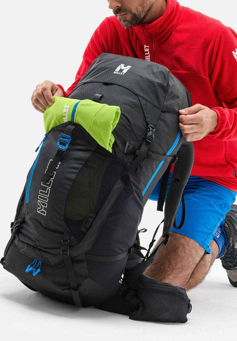 Schwarzer und blauer Millet-Rucksack mit einem oberen Fach und einer seitlichen Netztasche. Ein grüner Fleece wird in die Tasche gelegt. Verstellbare Träger sind sichtbar.