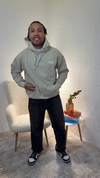 Beige hoodie with a front pocket and drawstring, dark jeans, and black sneakers with white accents. Standing in a softly lit room.