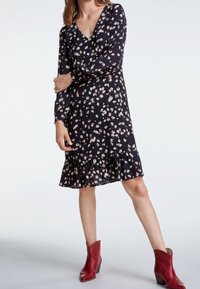 Robe noire à fleurs avec des manches longues, un décolleté en V et un ourlet volanté. Présente de petites fleurs roses et blanches sur un fond sombre.