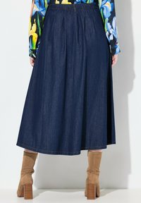 Jupe midi en denim bleu foncé, présentant des plis, une texture lisse et un ourlet droit. Associée à des bottines beige clair à talons empilés.