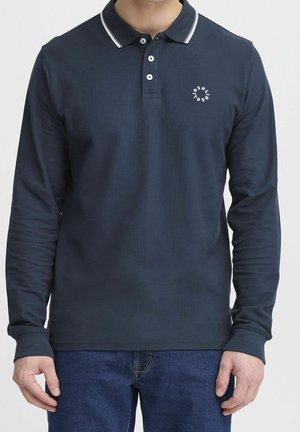Hombre que lleva una camisa polo azul marino de manga larga con logo y ribete blancos, combinada con vaqueros azules, de pie frente a un fondo liso.