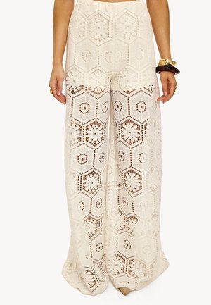 Mujer que lleva pantalones blancos de crochet de cintura alta y pierna ancha con patrones florales hexagonales, combinados con pulseras doradas y oscuras en la muñeca derecha.