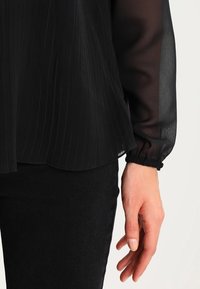 Svart plisserad blus med genomskinliga långa ärmar och klockade ärmslut, bärs över svarta byxor, som visar en hand som vilar mot tyget.