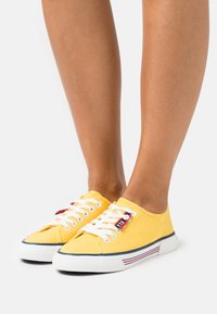 Żółte płócienne trampki z białymi sznurówkami, gumowymi podeszwami i paskowanym detalem wzdłuż krawędzi. Gładka faktura, okrągły design noska.